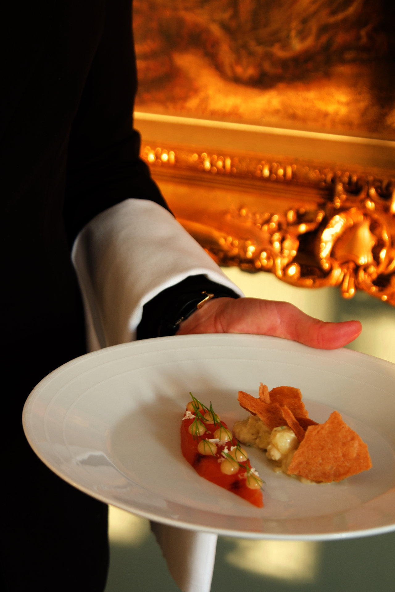 Hvit tallerken med liten rett anrettet, båret av en sortkledd person foran et maleri med gullramme.