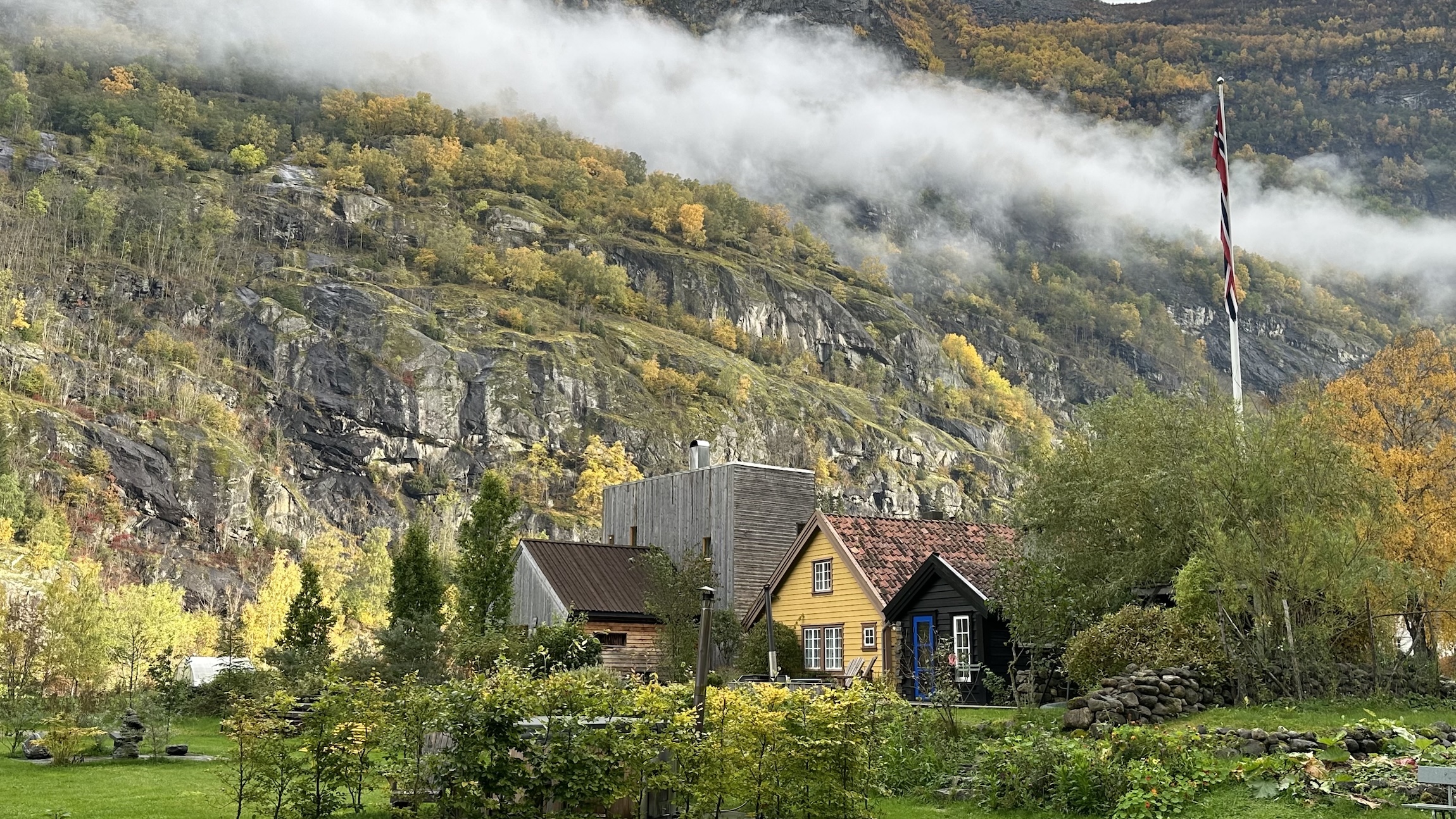 En klynge små tømmerhus på overnattingsstedet 292 Aurland omgitt av grønn mark og høy fjellvegg bak.